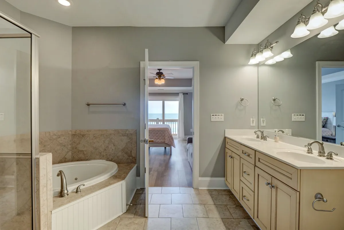 A luxurious bathroom with a soaking bathtub, dual sinks, a glass shower enclosure, and a doorway leading to a bedroom. The serene design is elevated by ample natural light and subtle views of the ocean, providing an ideal retreat for relaxation and self-care.
