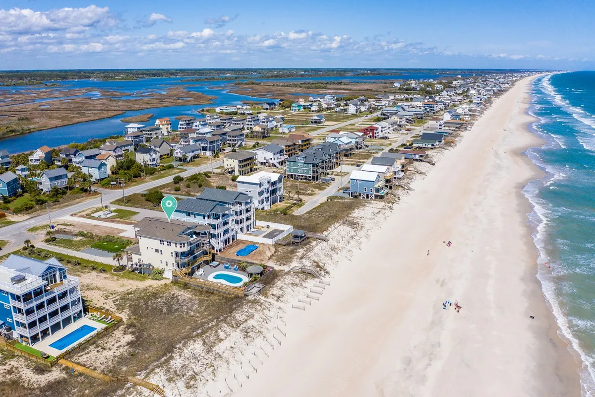This wide-angle view captures several beachfront properties lined up along the shore, each with inviting swimming pools, lush lawns, and the sparkling ocean just beyond. The coastline stretches into the distance, and the sandy beach is perfect for beachcombing, fishing, and water sports. The mood is relaxing and full of vacation promise.
