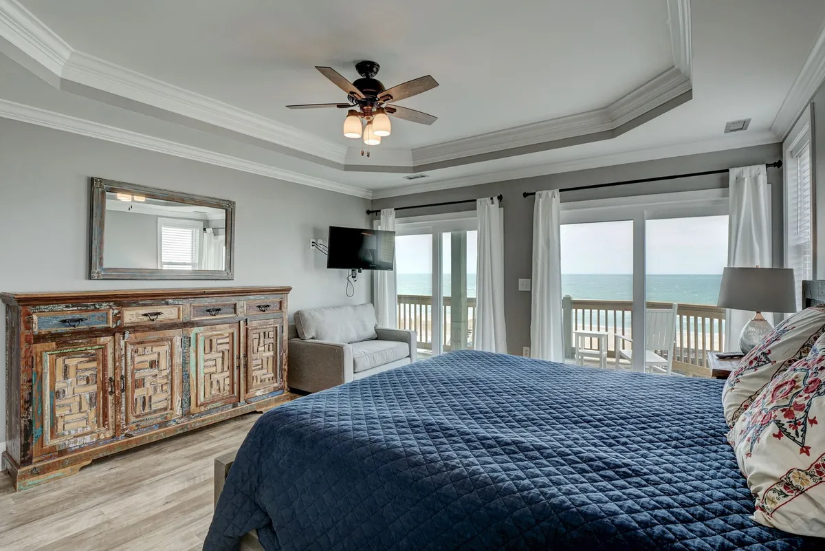 A cozy bedroom with a navy blue quilt on the bed, a rustic wooden dresser, large windows offering direct ocean views, and a comfortable couch. The space feels inviting and is ideal for relaxing, reading, or enjoying the scenery, whether alone or with a loved one (or a pet).