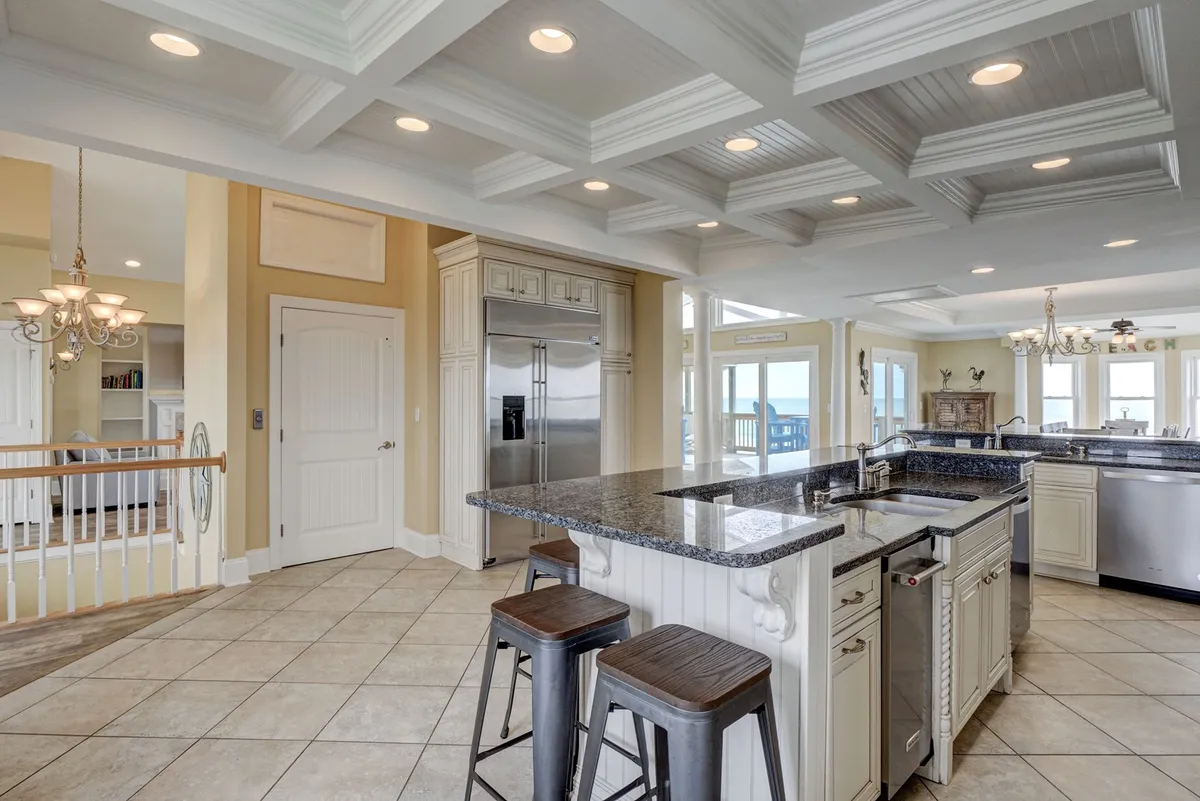 An elegant kitchen scene with a central island, bar stools, coffered ceiling, and large windows flooding the space with natural light. The kitchen looks open and connected to the dining area, designed for entertaining and gathering.