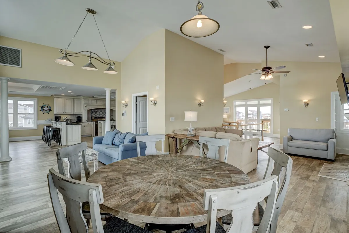 An inviting open-plan space combining a round wooden dining table, blue and beige sofas, and a visible kitchen area in the background. The scene exudes warmth and is clearly set up for family dinners, memorable gatherings, or lively game nights.