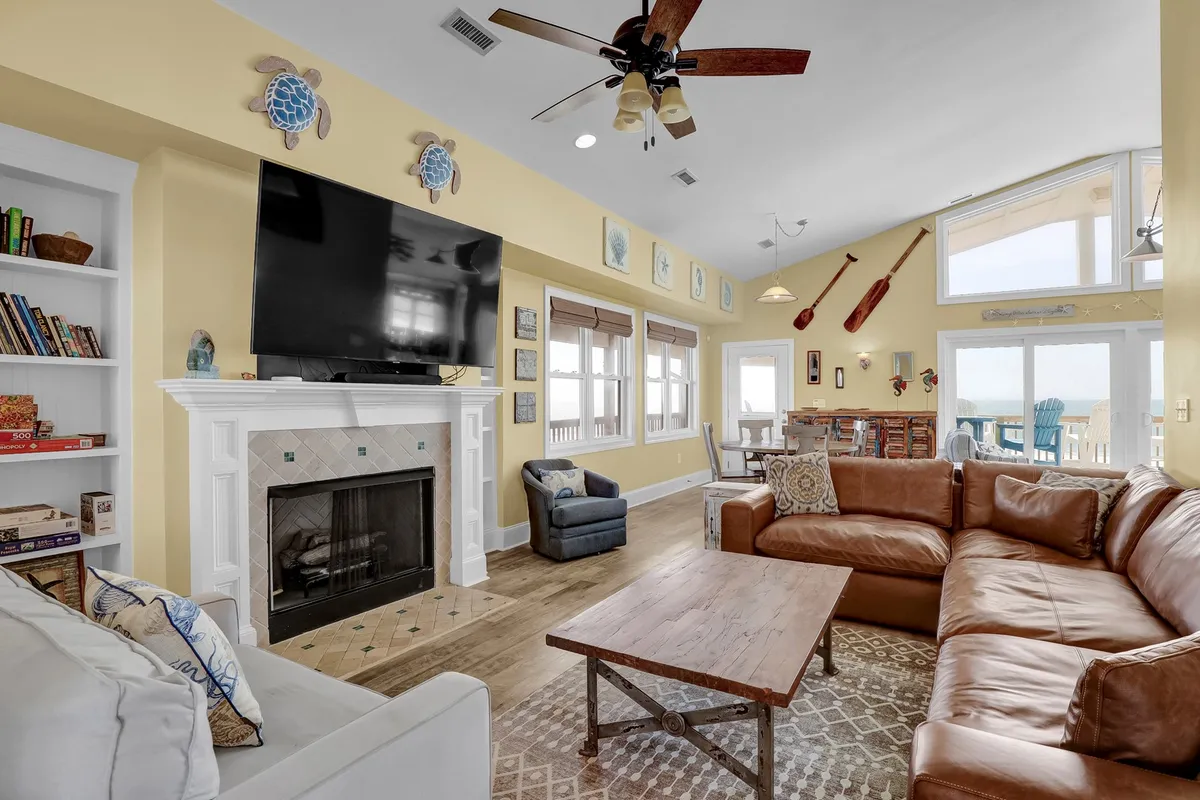 A spacious living area featuring a large sectional sofa arranged around a modern fireplace and TV, with natural light pouring in through generous windows that offer glimpses of the ocean. The hardwood floors gleam in the sunlight, giving the space warmth and elegance. The thoughtful arrangement invites groups to gather and relax together.