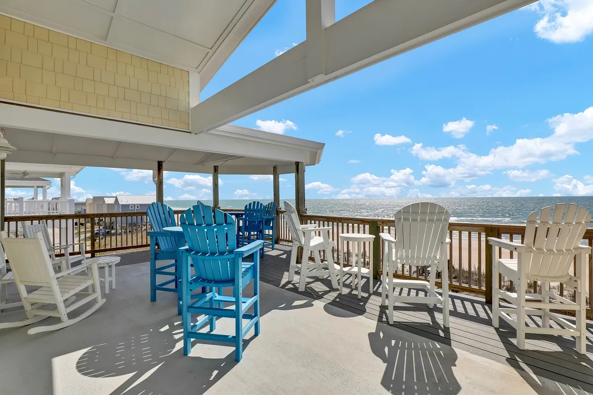 A deck furnished with blue and white Adirondack chairs overlooks a stunning ocean view, framed by a classic deck railing. The scene is tranquil, suggesting moments spent relaxing, sipping morning coffee, or gathering to watch the sunset over the waves.