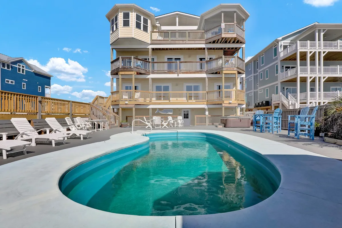 This image features a swimming pool in the foreground, with a multi-level house and balconies in the background. The deck area is equipped with chairs for lounging and there are ample spaces for social gatherings or barbecues. The mood suggests relaxed enjoyment with sunset views from the balconies.
