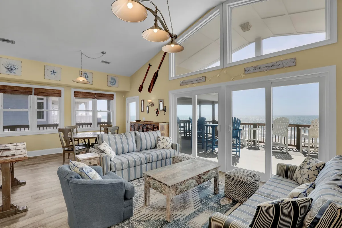 A vibrant living area with striped sofas and a rustic coffee table positioned in front of large windows that frame breathtaking ocean vistas. The room is filled with natural light, creating a cheerful and inviting atmosphere ideal for socializing and relaxing as a group.