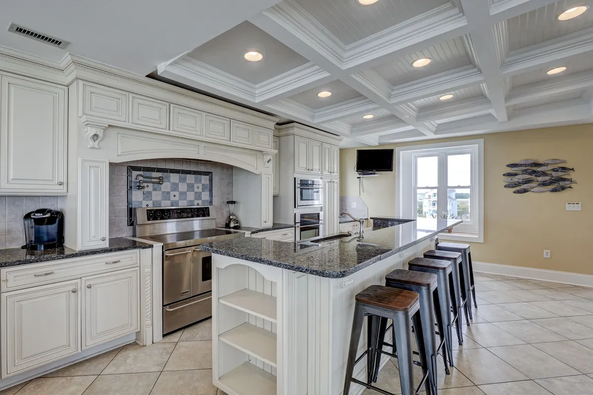 A spacious, welcoming kitchen featuring a granite countertop, stainless steel appliances, a coffered ceiling, and bar stools. The room is arranged for both cooking and socializing, with ample space for multiple people to gather and prepare meals together.