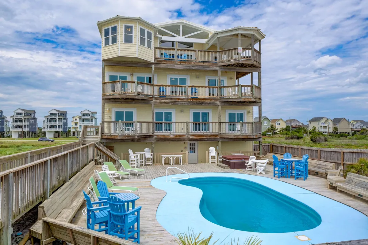 The image showcases a spacious, multi-story beach house with several balconies overlooking a sparkling swimming pool. Bright, colorful outdoor furniture invites relaxation, and the lush coastal landscape sets a tranquil backdrop. The scene radiates warmth, with spaces clearly designed for group gatherings, sunbathing, and outdoor dining.