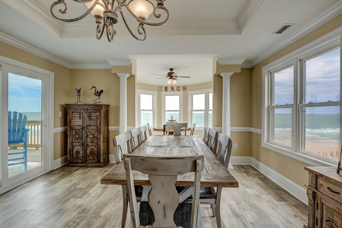 A large dining table set near expansive windows offers unobstructed ocean views. A stylish chandelier and beach-themed decor contribute to a serene and elegant atmosphere, ideal for group meals and celebrations.