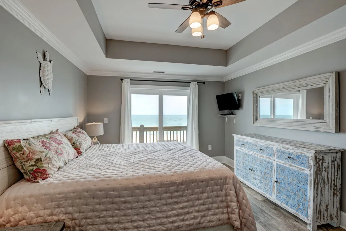 A tranquil bedroom featuring a bed adorned with floral pillows, a large window framing an ocean view, a decorative turtle on the wall, a blue and white dresser, and a ceiling fan with lights. Natural light streams in, creating a serene and inviting atmosphere perfect for restful nights.